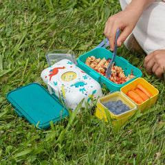 Bunte Kinder-Lunchbox mit Dino-Motiv, gefüllt mit Nudeln, Karotten und Heidelbeeren, auf Gras