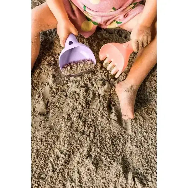 Kleinkind sitzt im Sand mit lila Schaufel und rosa Sandspielzeug, spielt barfuß im Sandkasten